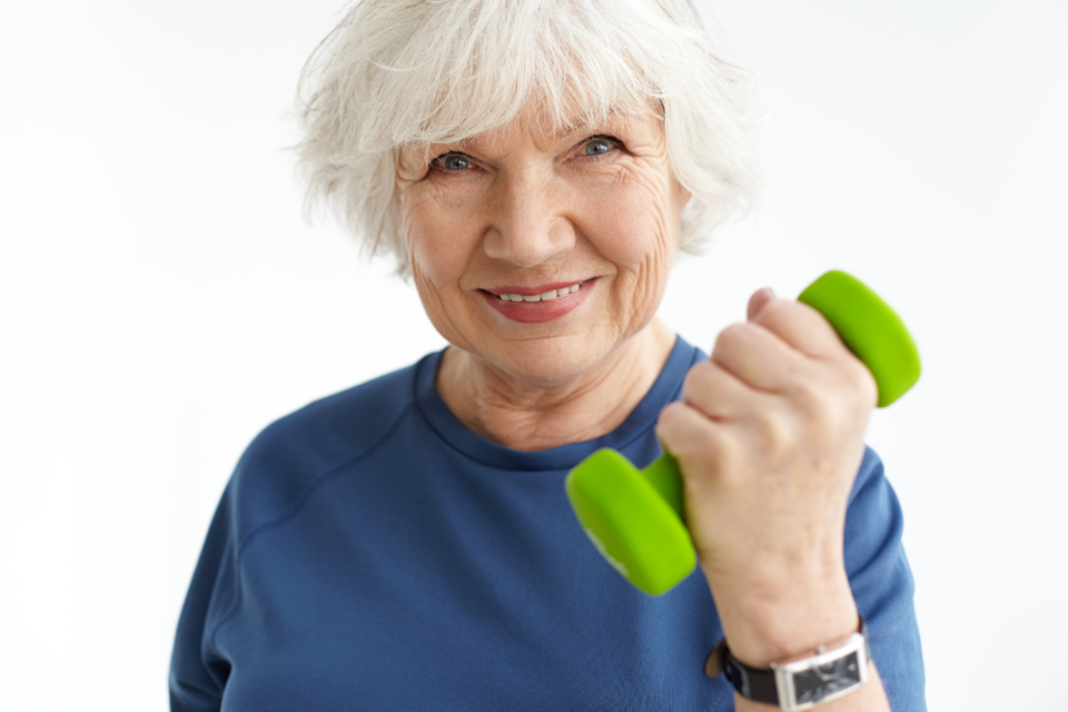 Close up image of sporty mature woman with gray hair doing bicep curls, holding green dumbbell and smiling happily at camera.